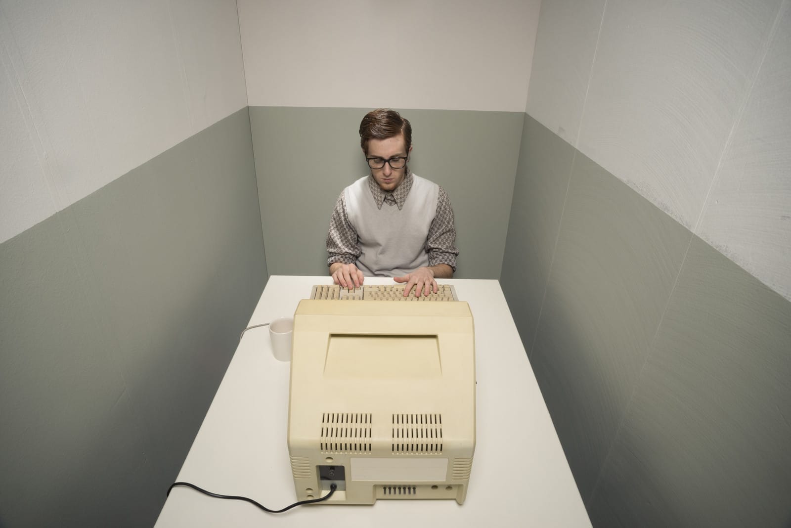 A person wearing glasses and a sweater vest sits at a small table in a narrow room, typing on a vintage computer with a large monitor. A mug is placed beside the computer. The walls are two-toned in light and dark gray.