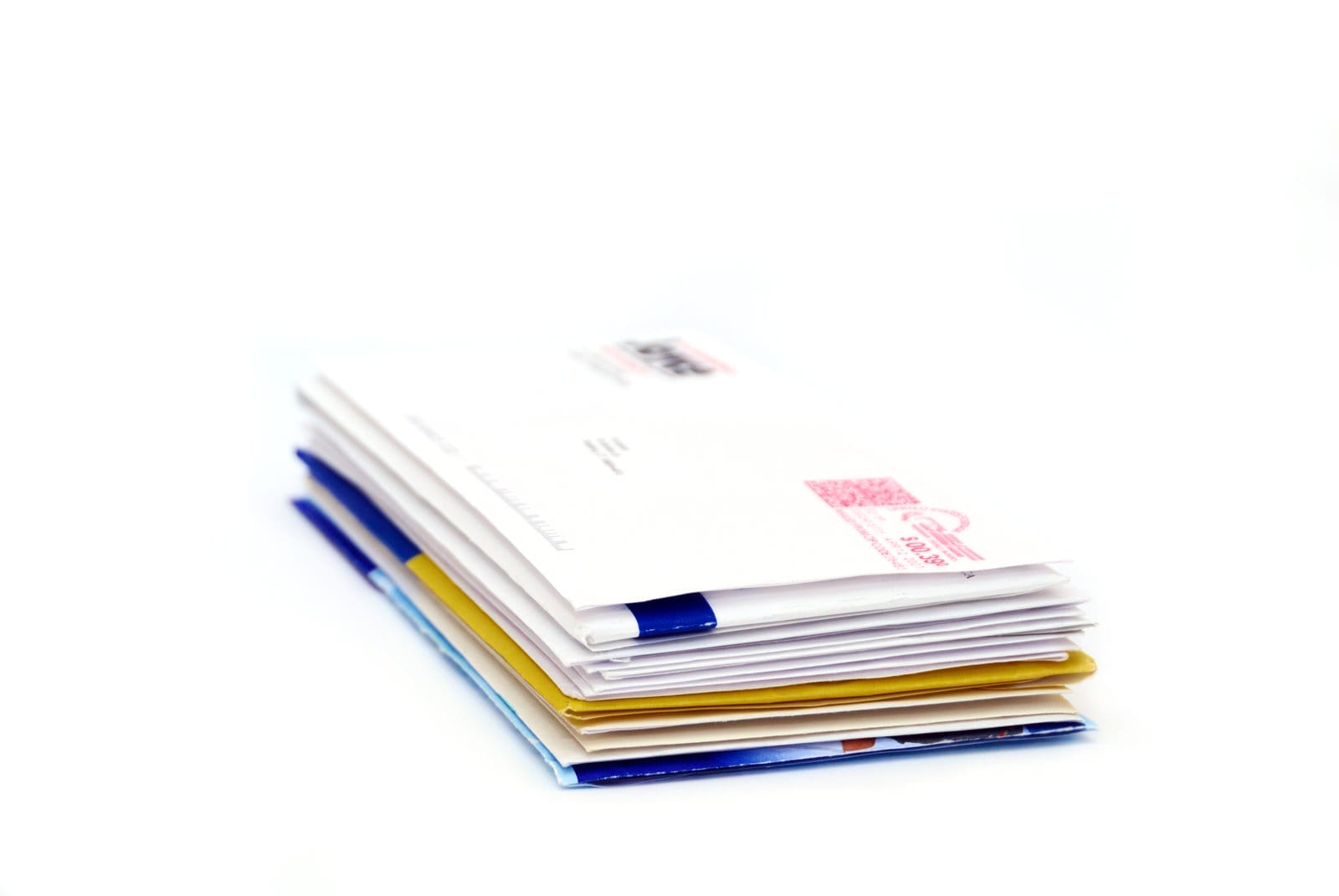 A stack of assorted envelopes lies on a flat surface. The envelopes vary in color and size, with some white, yellow, and blue. A visible postmark and stamp are on the top envelope. The background is plain white.
