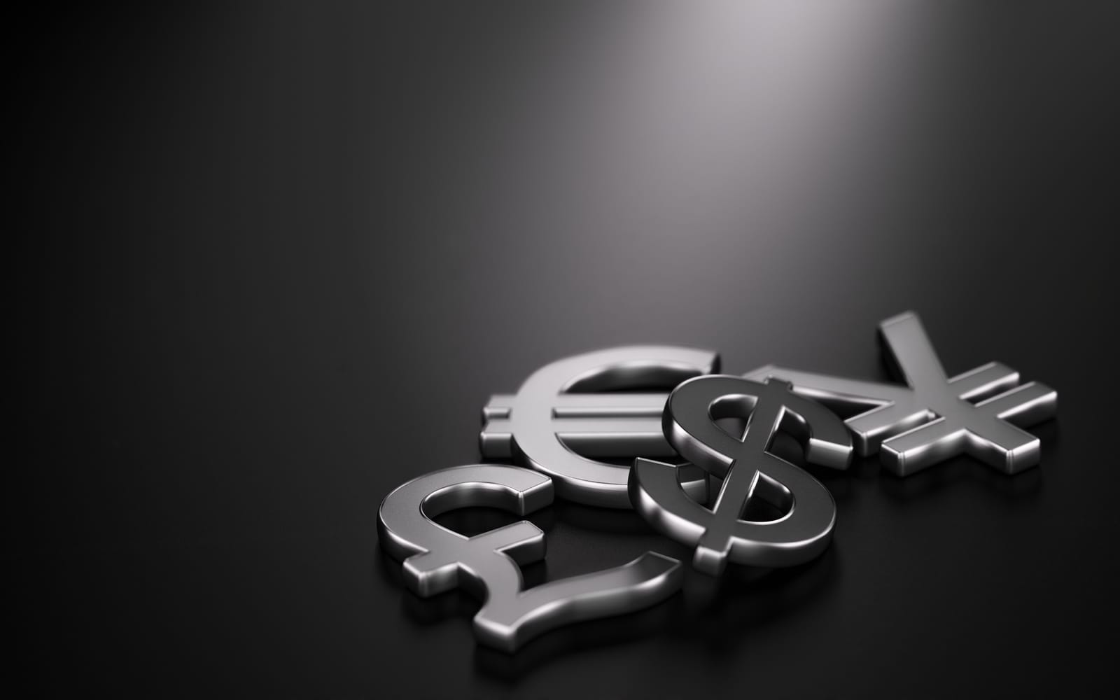 Silver currency symbols for the euro, dollar, yen, and pound lie scattered on a smooth, dark surface. The symbols are prominently placed with a soft spotlight highlighting their metallic texture.