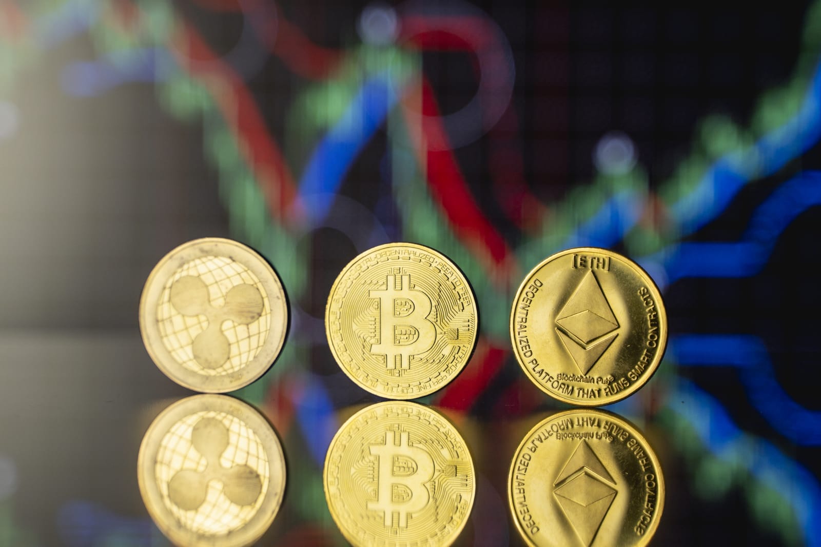 Three gold coins with cryptocurrency logos—Ripple, Bitcoin, Ethereum—are standing upright on a reflective surface. A digital screen displaying colorful financial charts is blurred in the background.