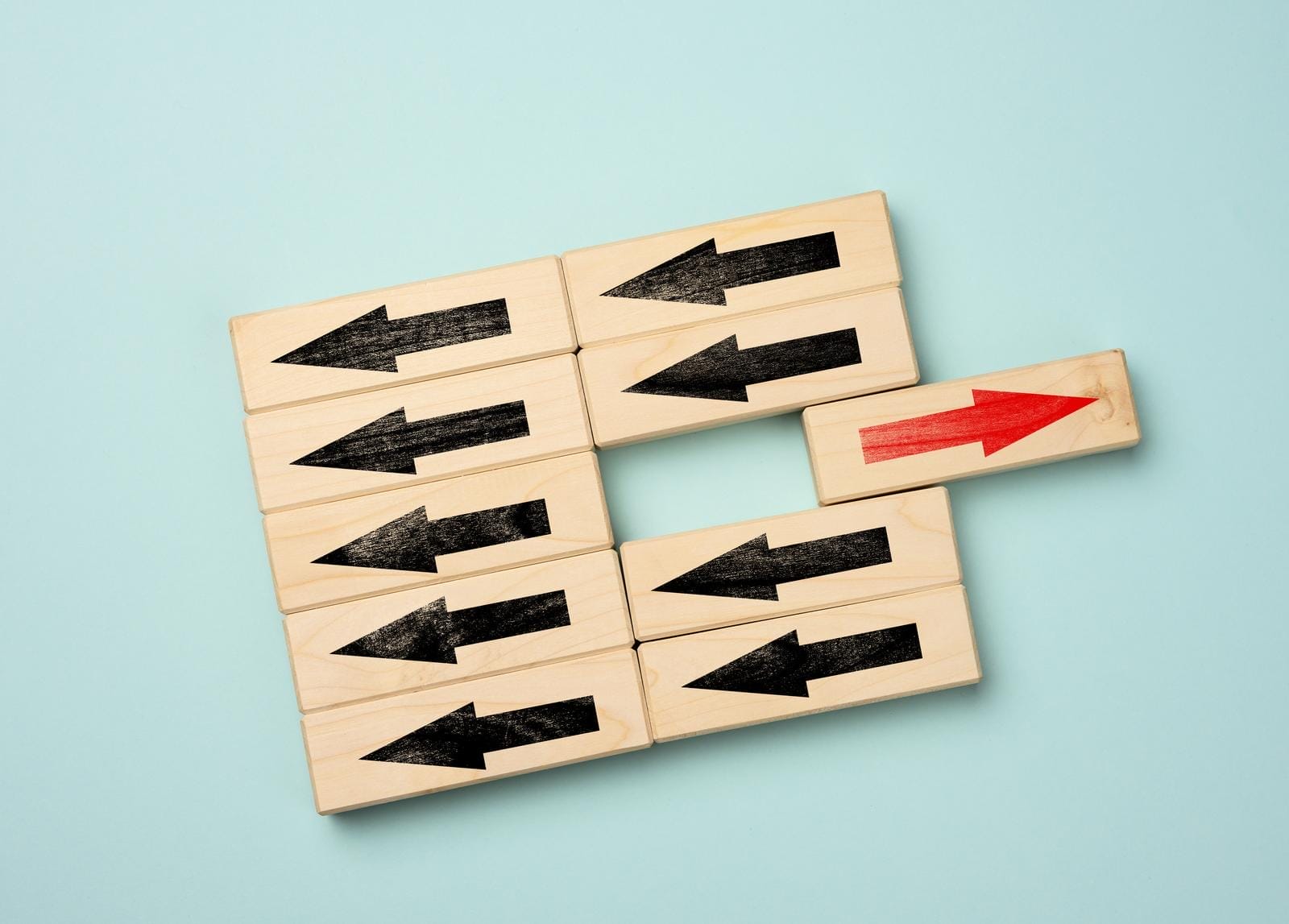 Wooden blocks with black arrows pointing left, arranged in a square shape, against a light blue background. One block with a red arrow points to the right, breaking the pattern and extending out from the square.