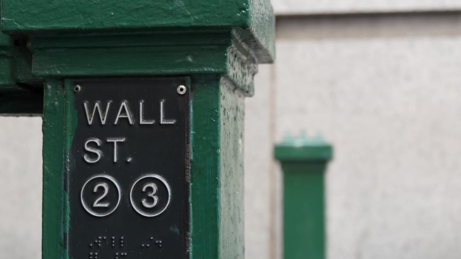 Close-up of a green metal post with a black sign reading "WALL ST." and symbols "2" and "3" indicating train lines. The background is blurred, with another green post partially visible.
