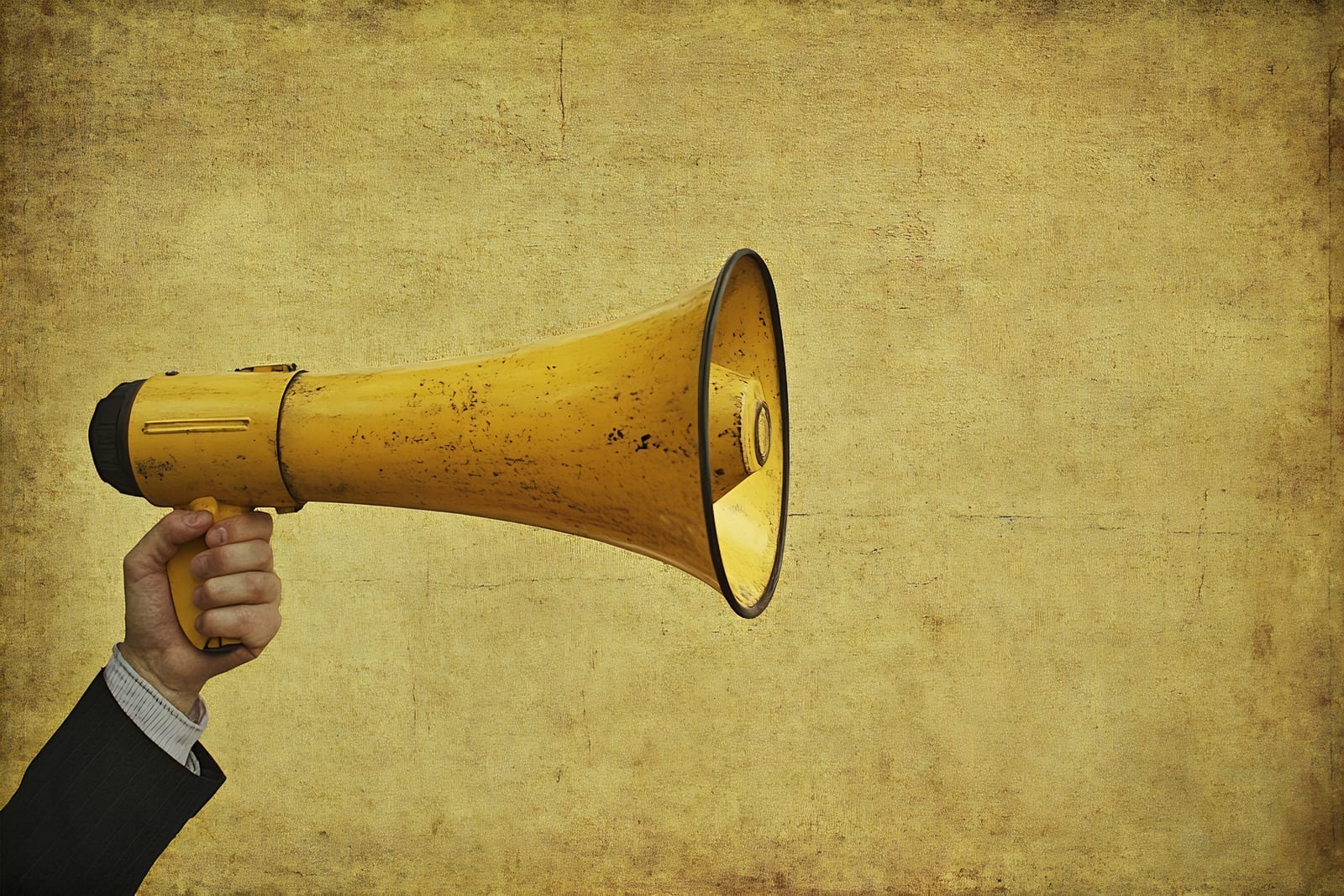 A person in a suit holds a yellow megaphone against a textured, grunge-style yellow background. The image focuses on the megaphone, which points to the right, with the person's hand emerging from the left.