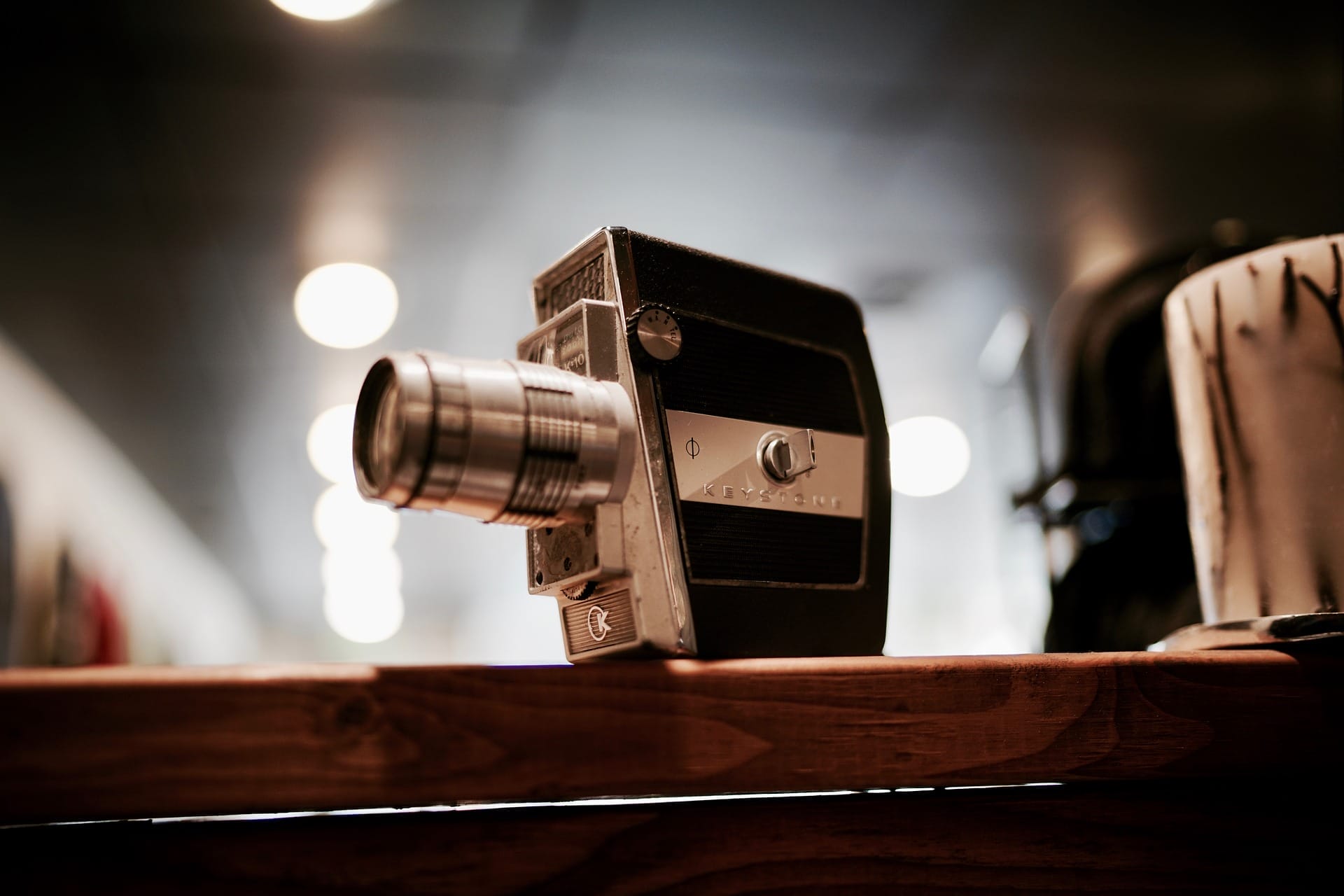 A vintage video camera with a large lens sits on a wooden surface. The background is softly blurred with several round lights creating a bokeh effect. The camera has a black and silver body with textured details.