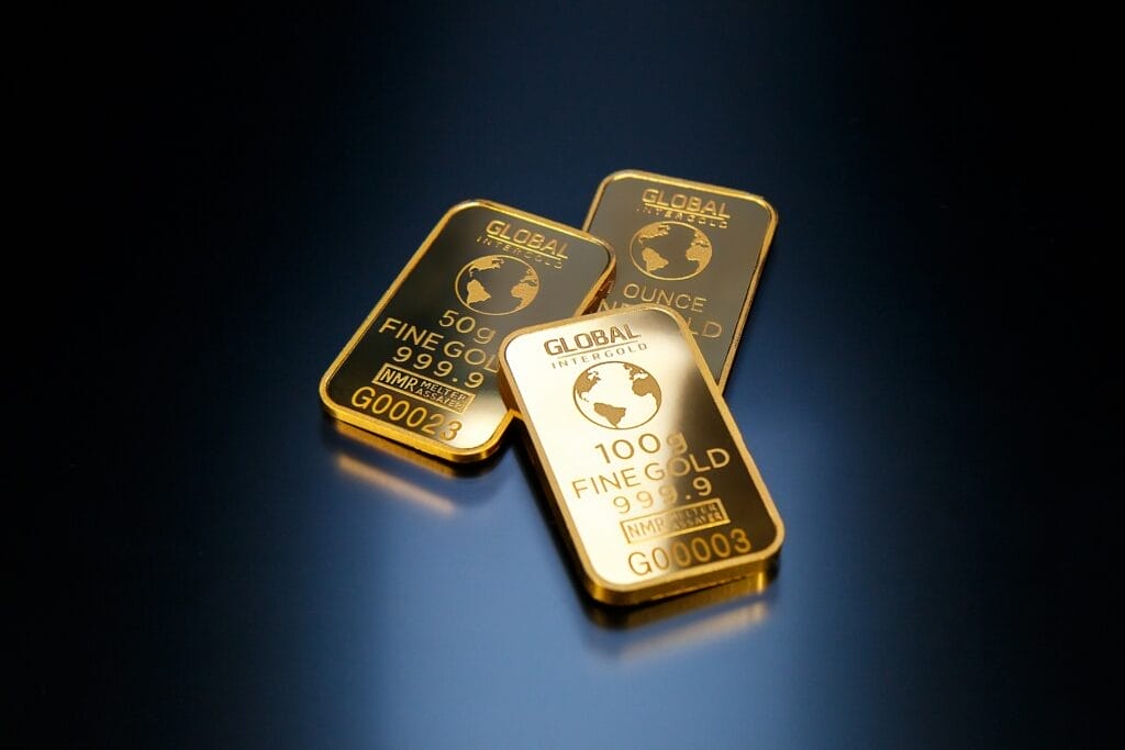 Three gold bars on a dark surface, each inscribed with "GLOBAL ITEC GOLD" and their respective weights: 50g, 100g, 1 ounce. The bars have a shiny finish, reflecting light.