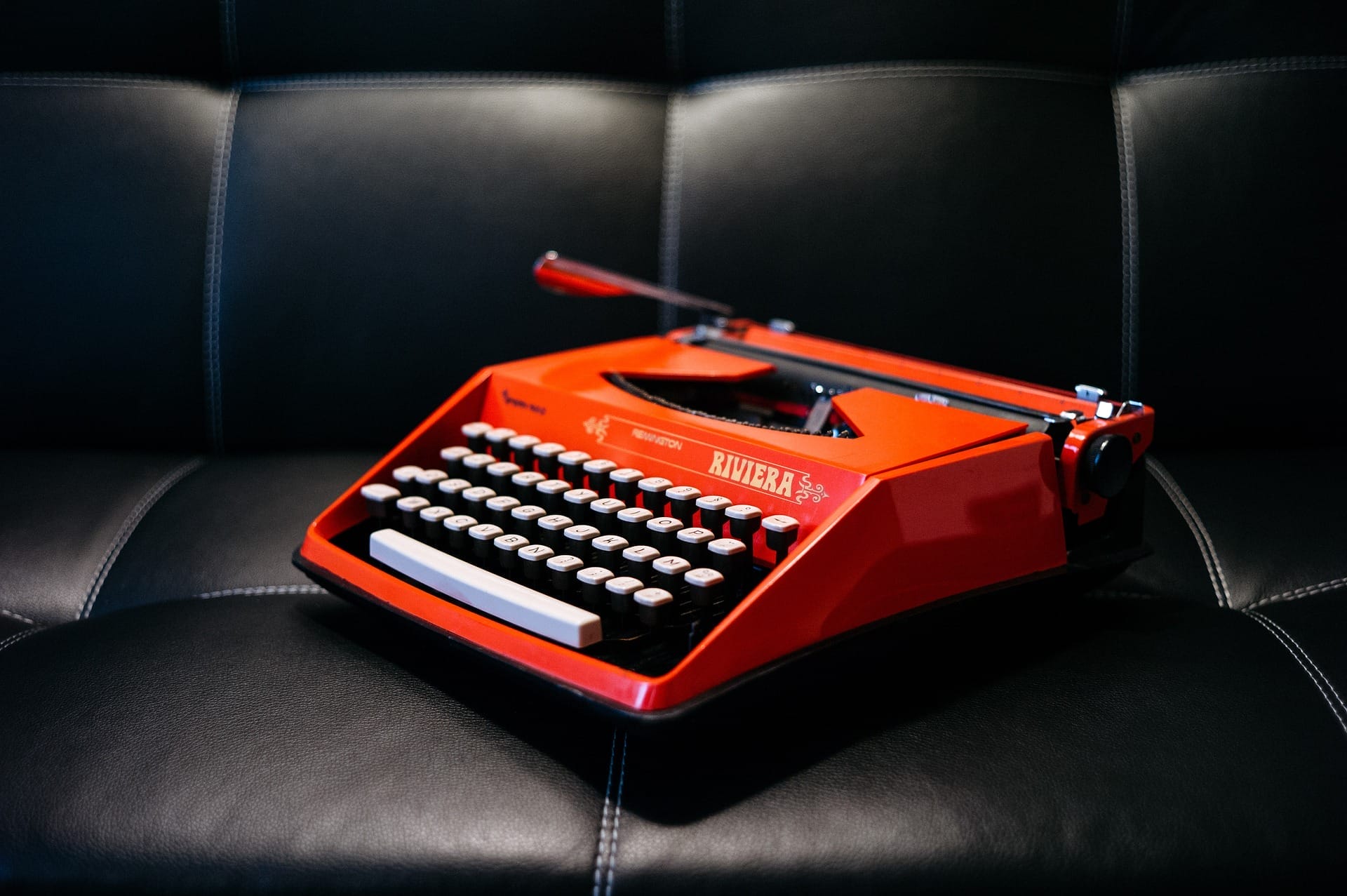A vintage red typewriter is placed on a black leather couch. The typewriter has white keys and chrome features, creating a striking contrast with the sleek surface of the couch.