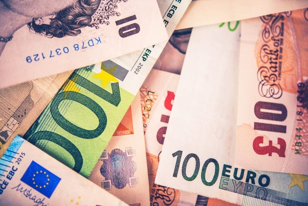 A pile of various international banknotes, including 100 and 10 Euro notes, a 10 British Pound note, and others, overlapping each other. The currencies display colorful designs, numbers, and symbols.