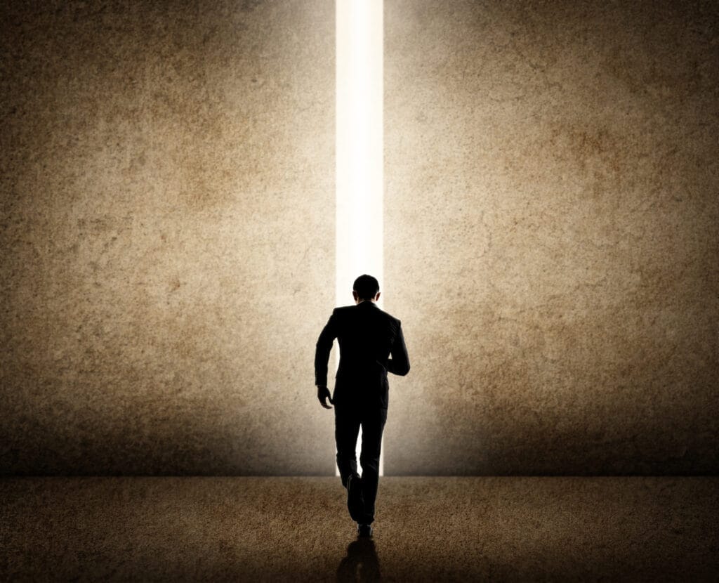 A person in a suit walks toward a narrow vertical beam of bright light shining through a crack in a large, textured wall, creating a dramatic and hopeful atmosphere.