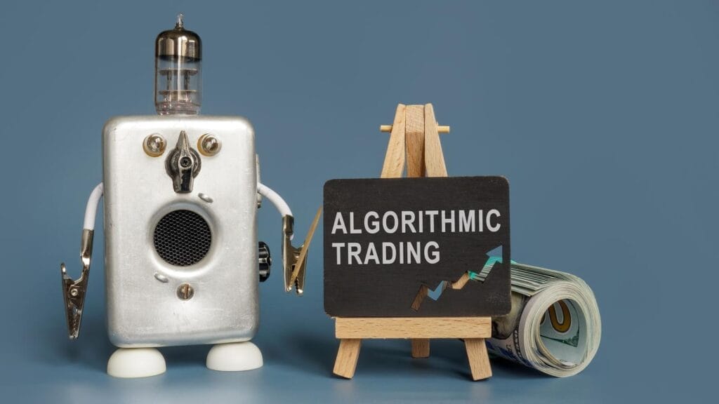 A small robot stands beside a chalkboard on an easel displaying the words "Algorithmic Trading" and an upward-pointing graph, with a roll of U.S. dollar bills next to it, against a plain blue background.