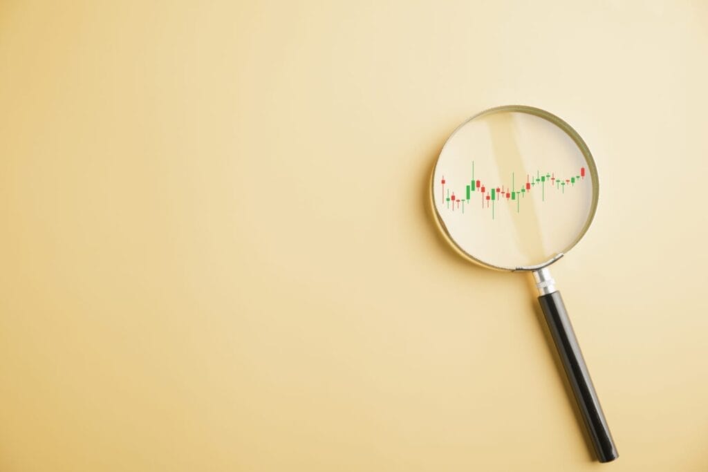 A magnifying glass lying on a beige surface, focusing on a candlestick stock chart with red and green bars visible inside the lens.