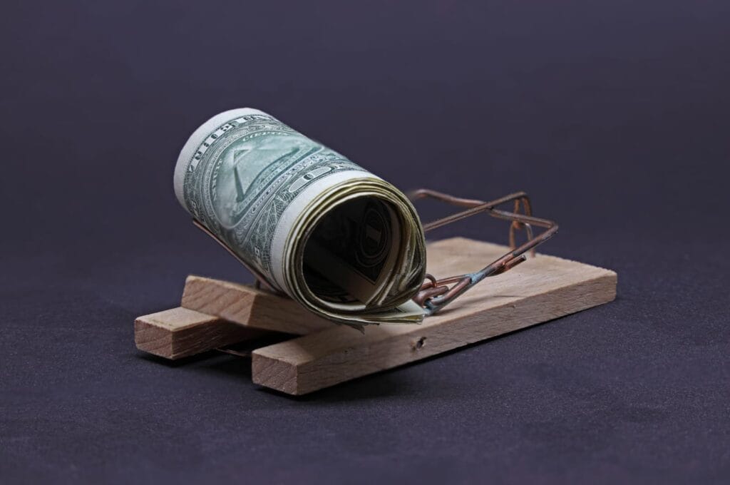 A rolled-up US dollar bill placed as bait on a wooden mousetrap, set against a dark background, symbolizing the dangers or risks associated with money.