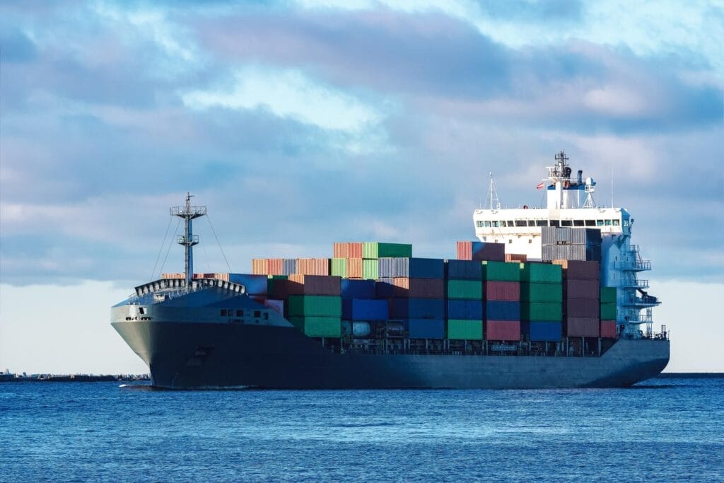 A large cargo ship loaded with colorful shipping containers sails across calm blue water under a partly cloudy sky.