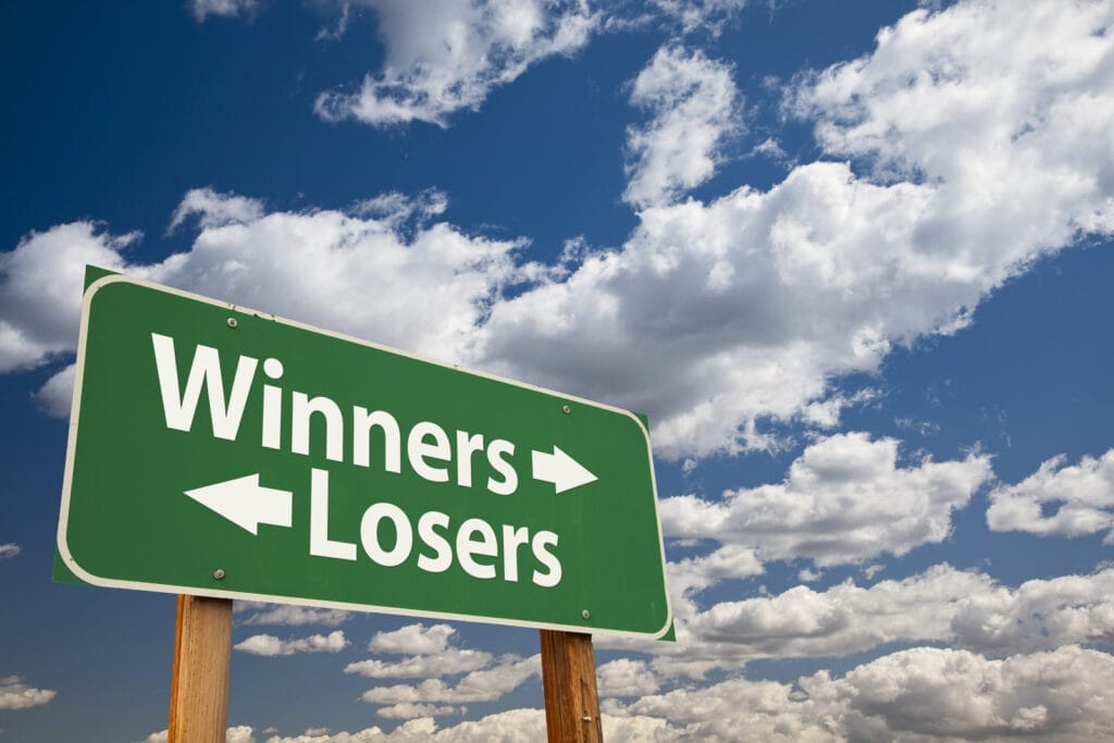 A green road sign with "Winners" and an arrow pointing left, and "Losers" with an arrow pointing right, stands against a bright blue sky with scattered clouds.